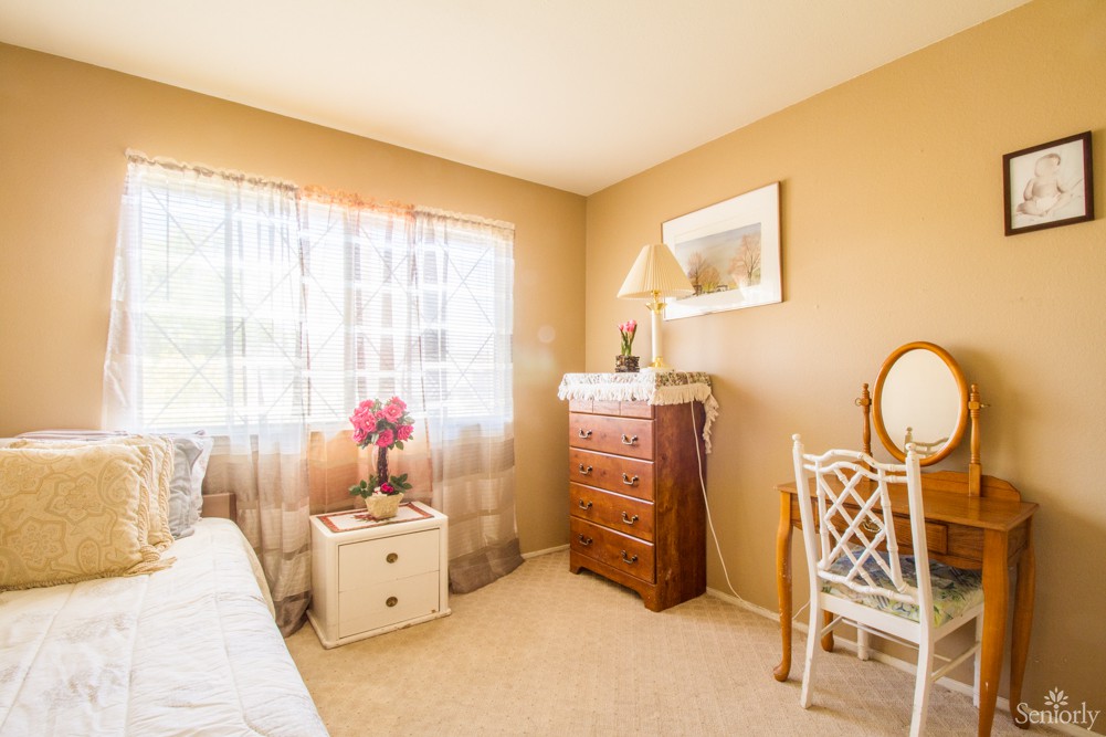 A cozy bedroom with natural light streaming through a window, featuring a dresser, chair, and decor.
