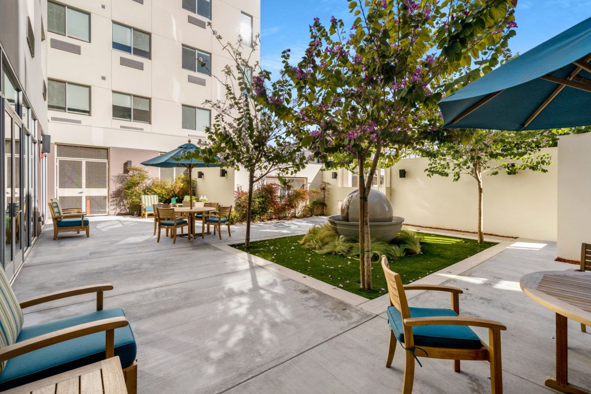 A serene courtyard with outdoor seating and umbrellas, surrounded by greenery and a multi-story building in the background.