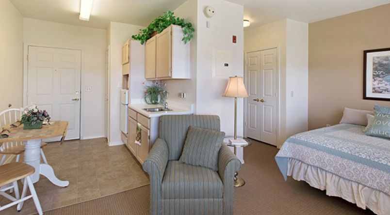 A cozy living space featuring a comfortable armchair, a kitchenette, and a neatly made bed.