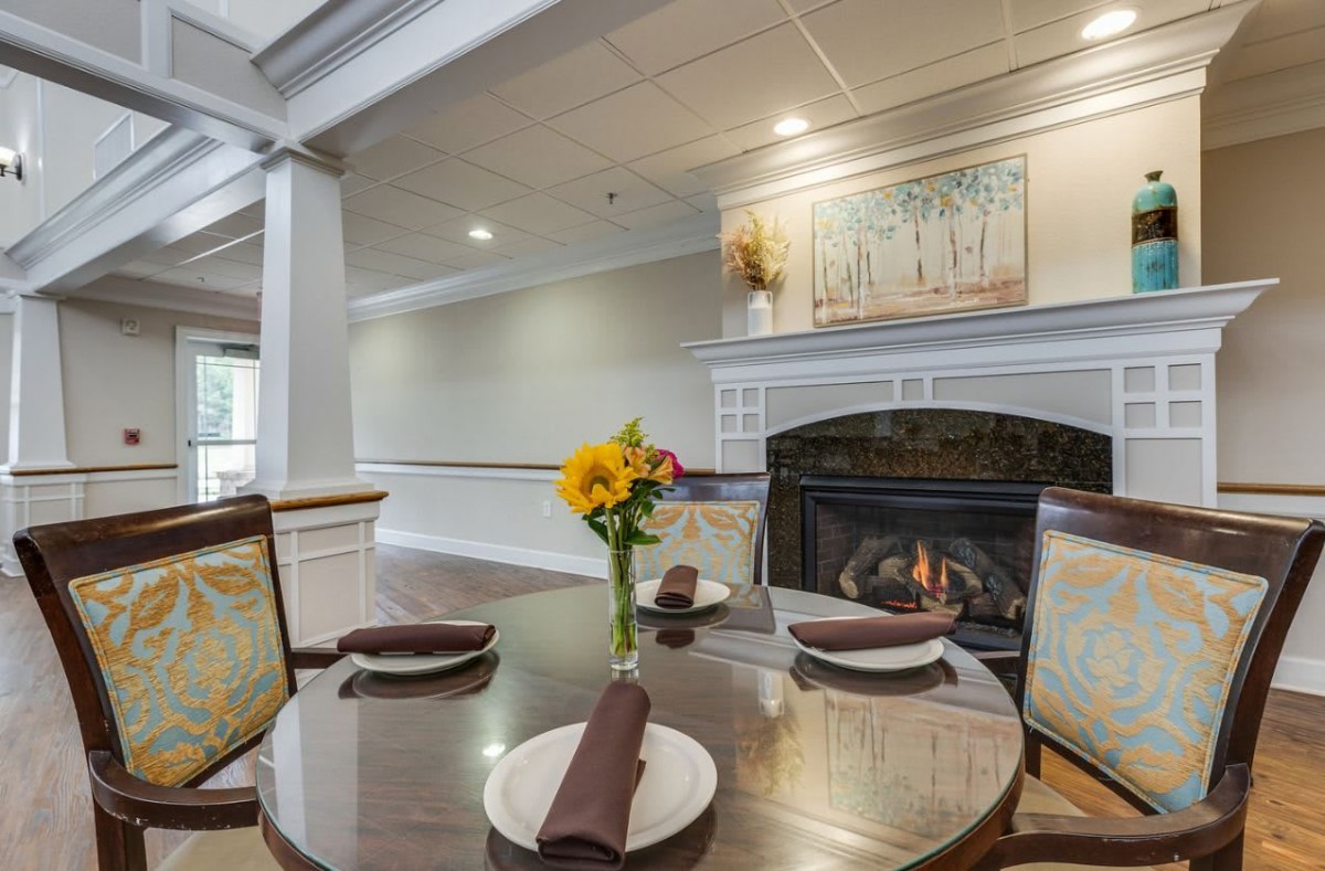 A cozy dining area featuring a table set for two with brown napkins, a bright flower vase centerpiece, and a warm fireplace in the background.