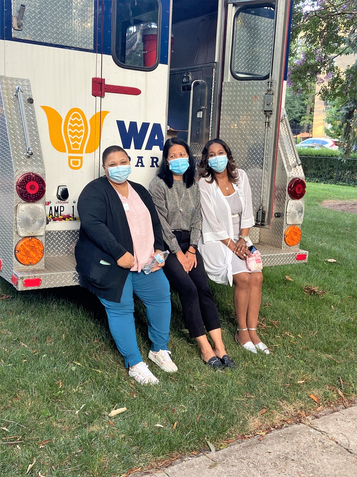 Three people wearing masks sit outside near a medical truck.