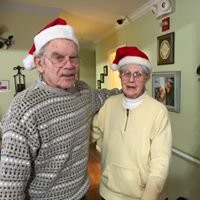 An elderly couple wearing Santa hats and sweaters, posing indoors during the holiday season.