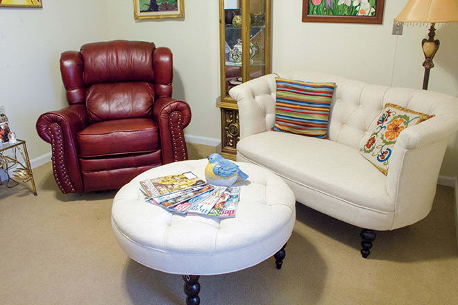A cozy living room features a red leather armchair, a tufted white sofa with colorful pillows, and a round ottoman displaying assorted magazines.