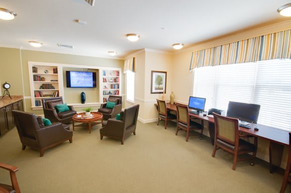 A cozy living room with a seating area, computer stations, and a television, surrounded by bookshelves and windows.