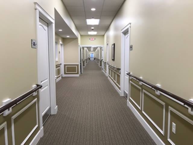 A long, well-lit hallway with carpeted floors, lined with doors and handrails.