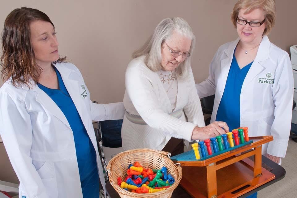 An elderly woman engages in a therapeutic activity with the assistance of two healthcare professionals.