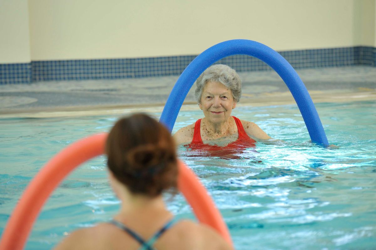 An elderly woman participates in a water aerobics class using pool noodle floats.