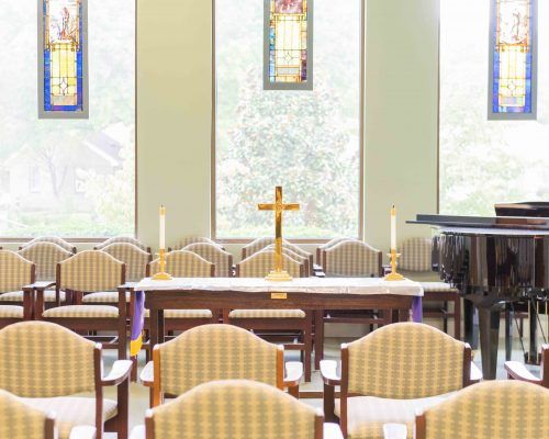 A peaceful chapel interior featuring an altar with a cross, surrounded by seating and stained glass windows.