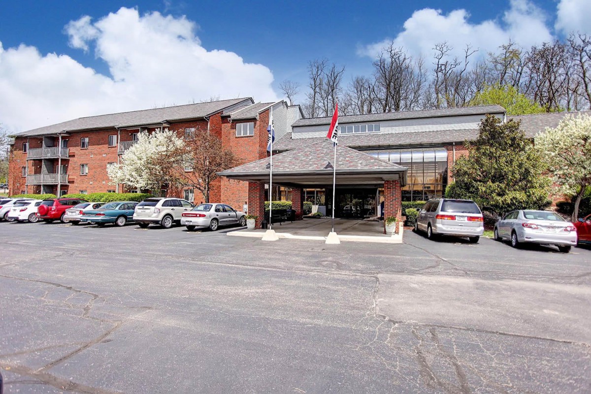 A building with a parking lot and several cars, surrounded by trees under a partly cloudy sky.