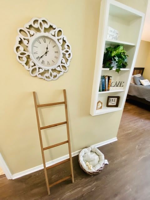 A cozy room corner featuring a decorative wall clock, a small wooden ladder, and a built-in bookshelf with plants and books.