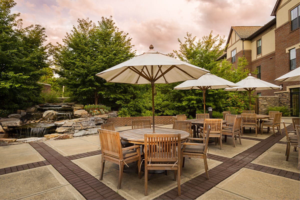 A tranquil outdoor patio area with wooden tables and chairs under large umbrellas, surrounded by lush greenery and set against a warm sunset sky.