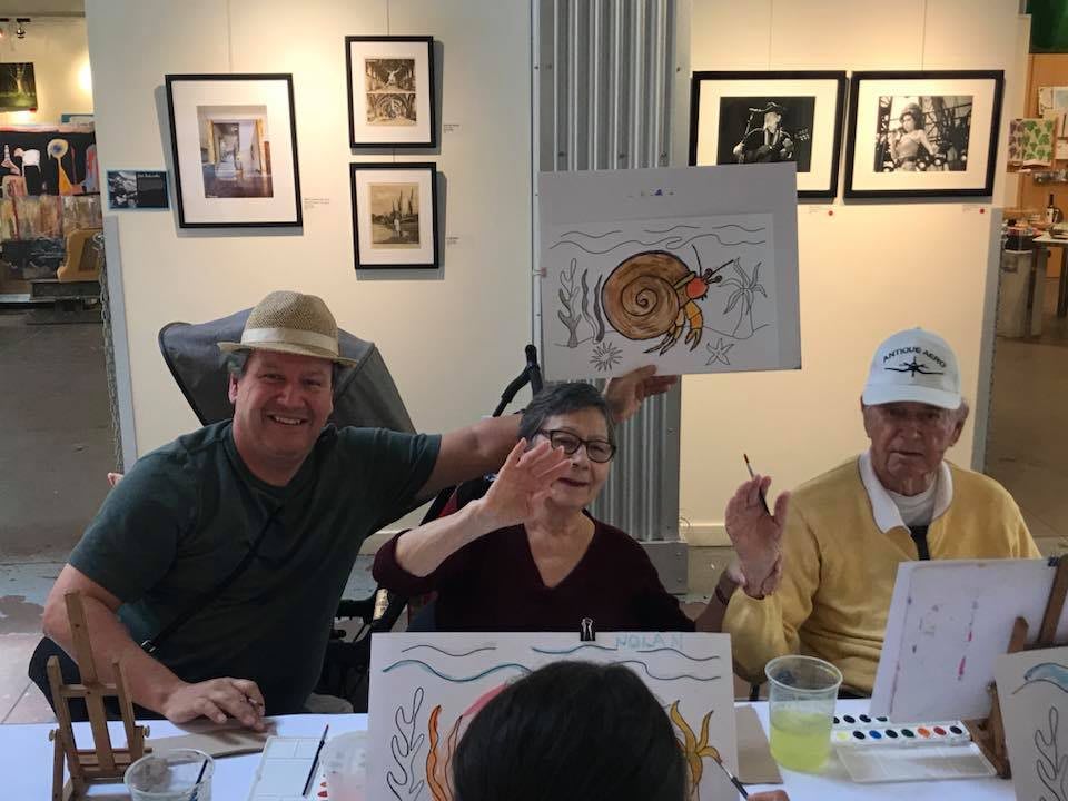 A group of people enjoying an art event, holding their drawings and smiling at the camera.