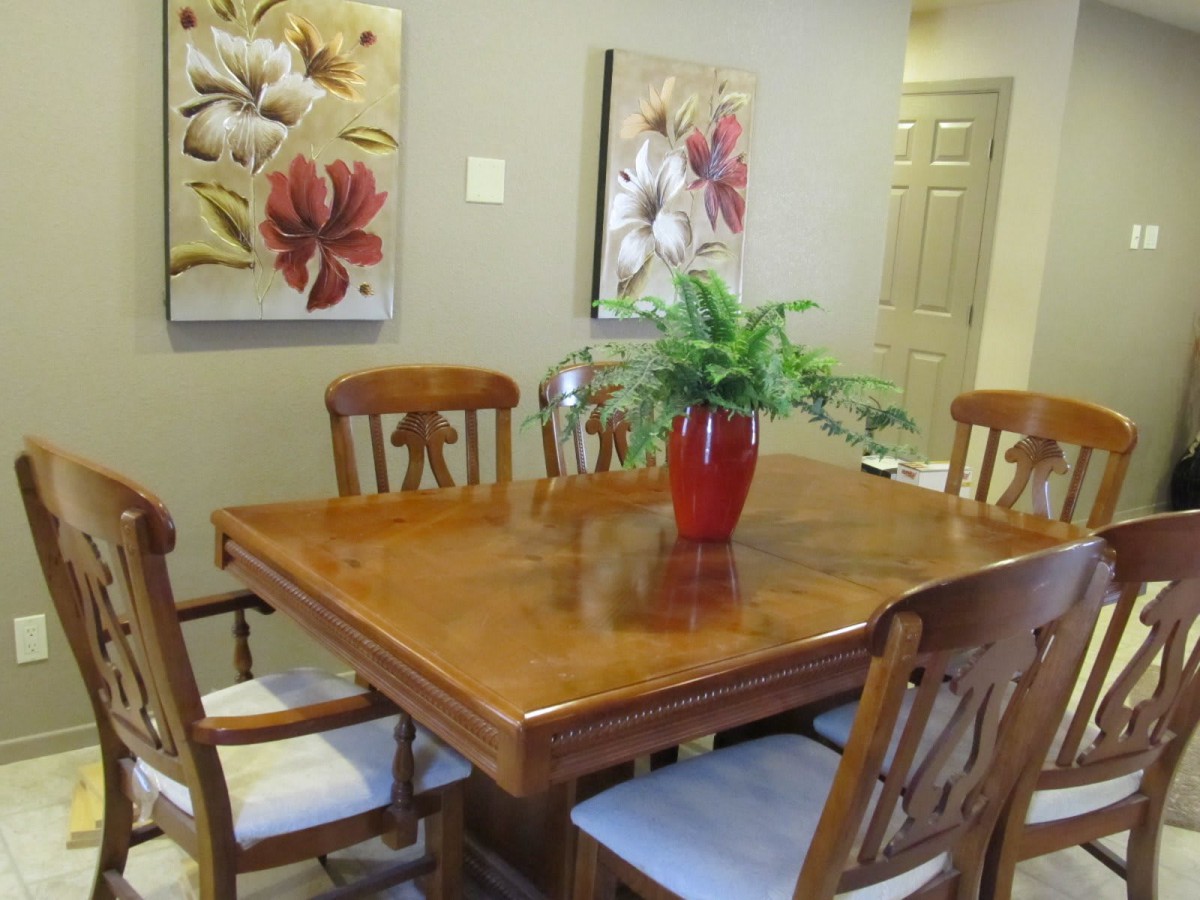 A cozy dining room with a wooden table, surrounded by chairs, adorned with a red vase and greenery. Floral paintings hang on the wall.