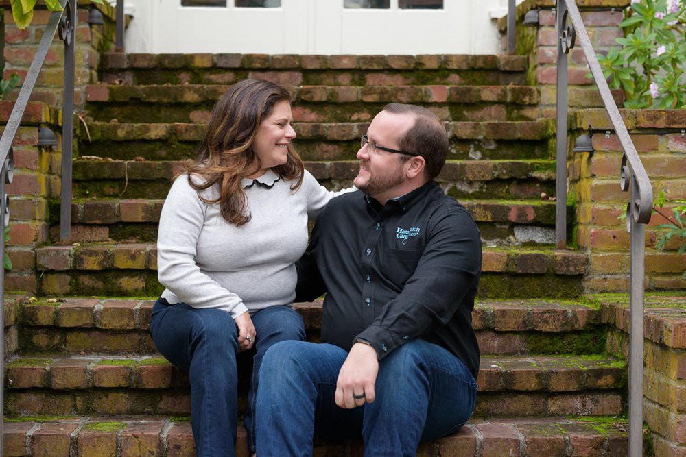 A couple sitting on outdoor stairs, smiling at each other in casual attire.