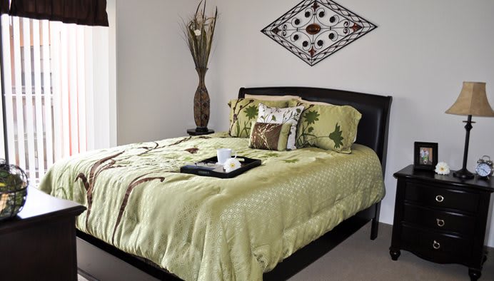 A cozy bedroom with a neatly made bed featuring green bedding and decorative pillows, complemented by wall art and a vase in the corner.