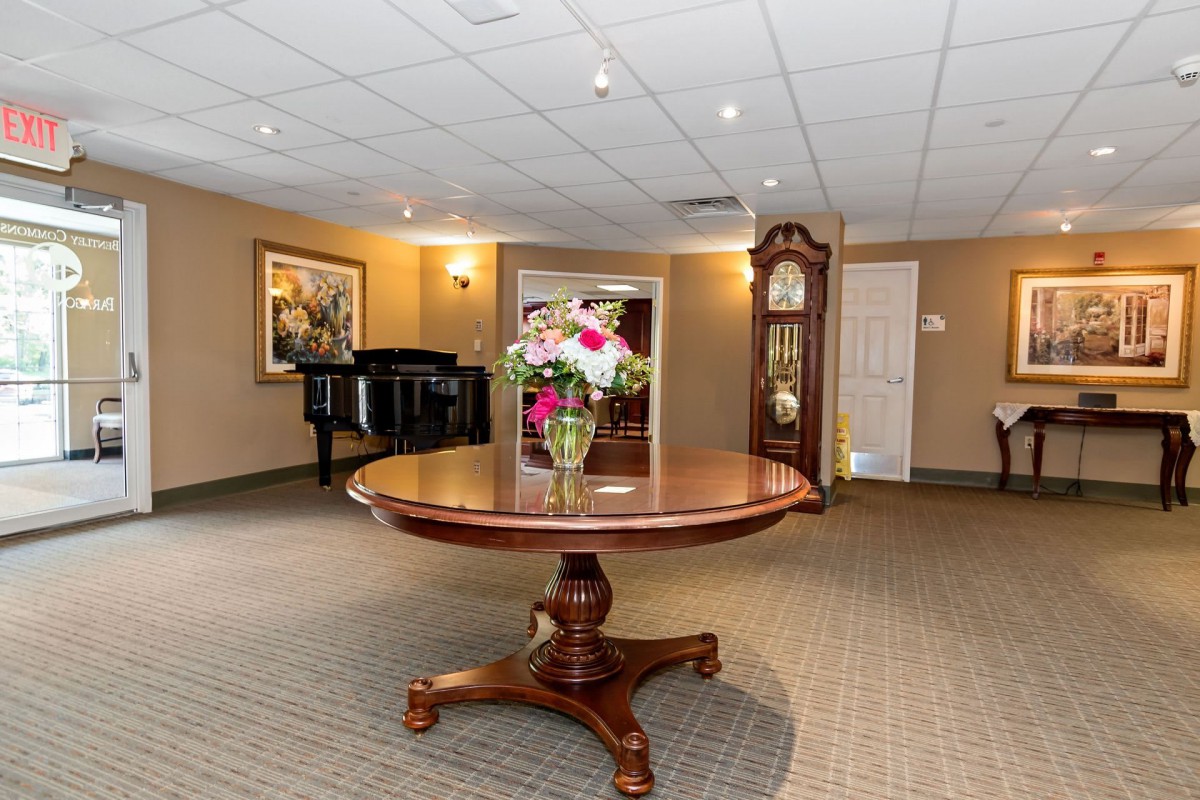 A spacious room featuring a round wooden table with a flower arrangement, a grandfather clock, and a piano in the corner.