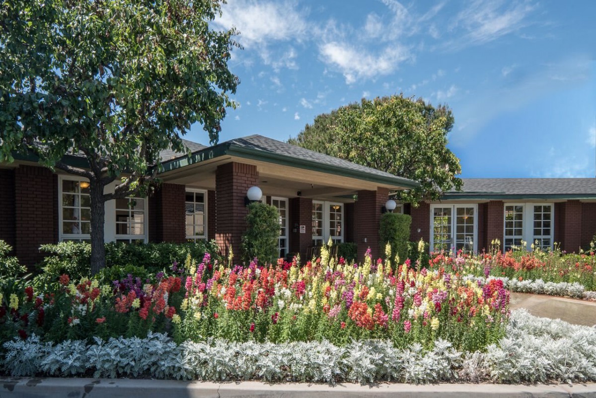 A charming house surrounded by a colorful garden with vibrant flowers and lush trees under a blue sky.