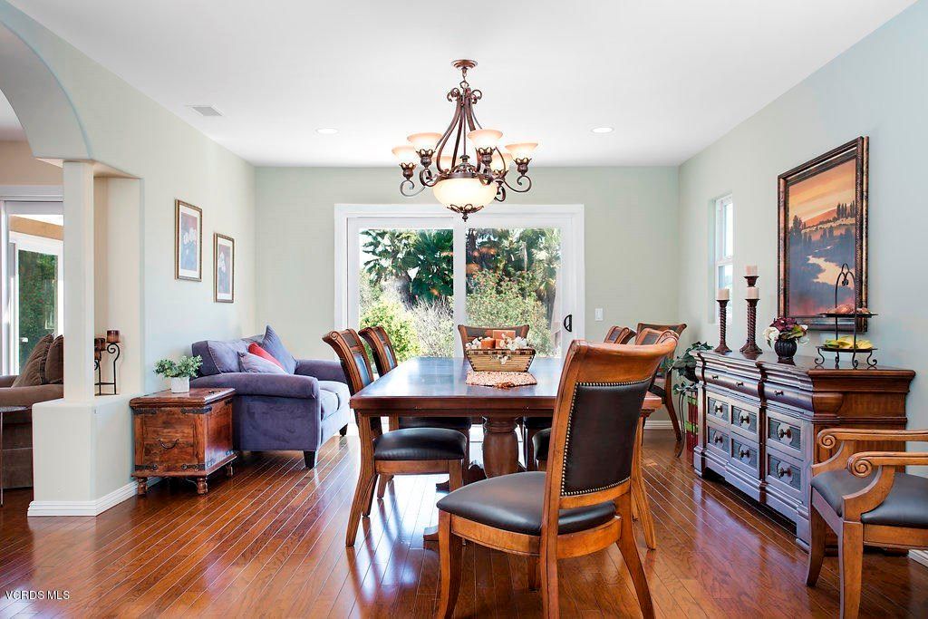 A well-lit dining room with a wooden table and chairs, featuring a chandelier and natural light from large windows.