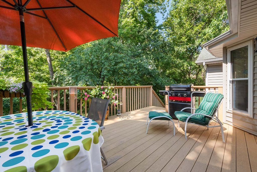 A sunny deck with a table, umbrella, lounge chairs, and a grill surrounded by trees.