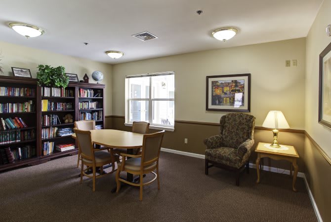 A cozy library room with a table and chairs, bookshelf, and window lighting.