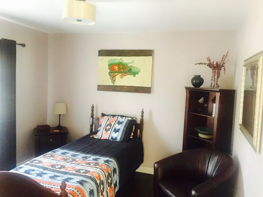 A cozy bedroom with a single bed, adorned with patterned bedding, a wall painting, and a nearby bookshelf and chair.