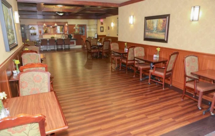 A cozy restaurant dining area with wooden floors, tables, and chairs, softly lit by wall lights.