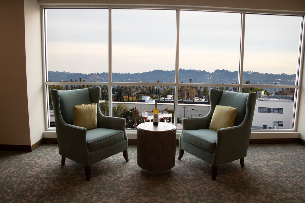 Two armchairs and a table with a bottle set against a scenic mountain view through large windows.