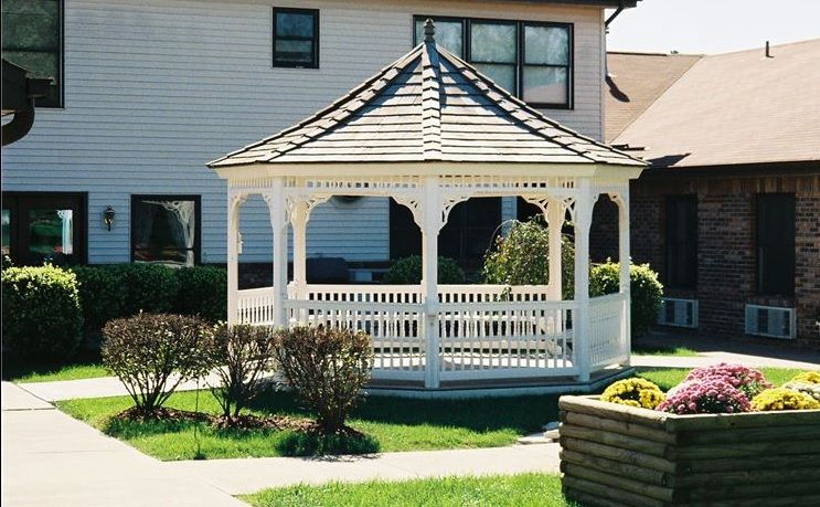 A wooden gazebo situated in a sunny yard with manicured lawn and shrubs.