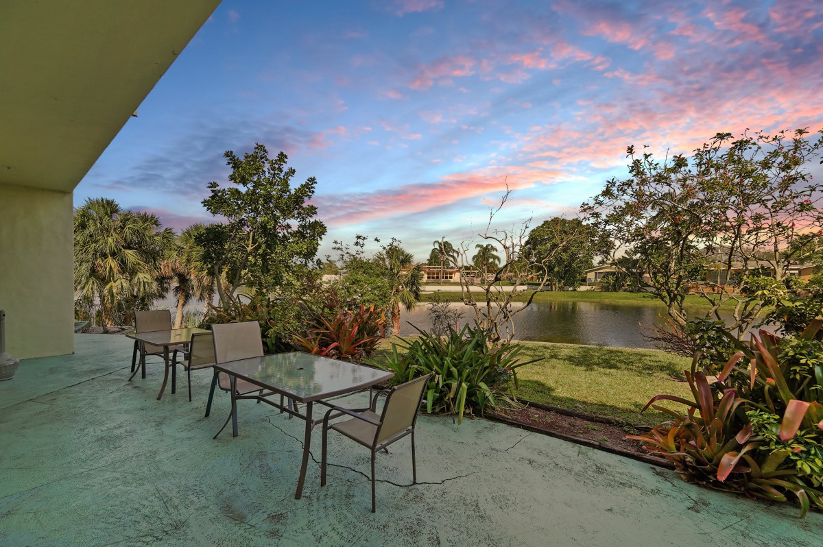 A scenic patio view with chairs and tables overlooking a garden and lake at sunset.