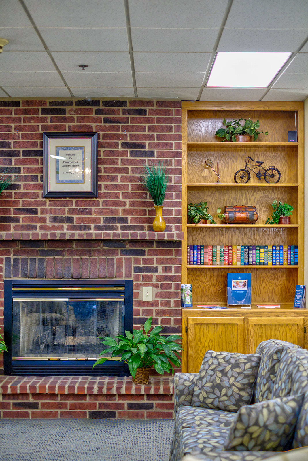 A cozy living room with a brick fireplace, wooden bookshelf, and decorative plants.