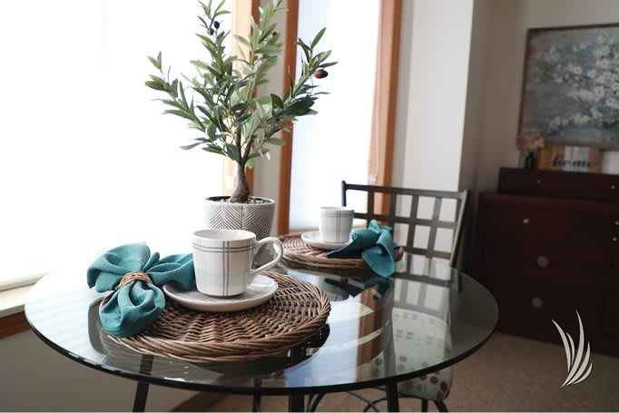 A cozy dining setup with a glass table featuring white tea cups, blue napkins, and a potted plant centerpiece.