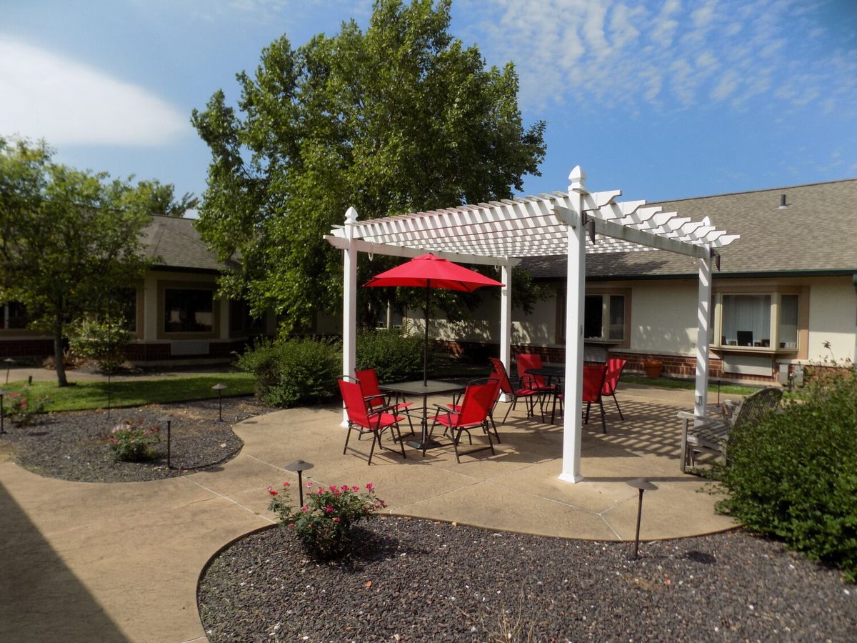 A cozy outdoor patio with red chairs under a white pergola and umbrella on a sunny day.