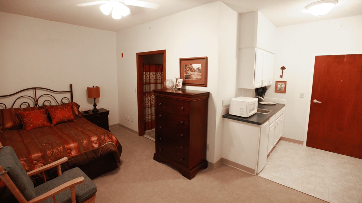 A cozy bedroom with adjoining kitchen area, featuring a bed, chest of drawers, and a microwave.