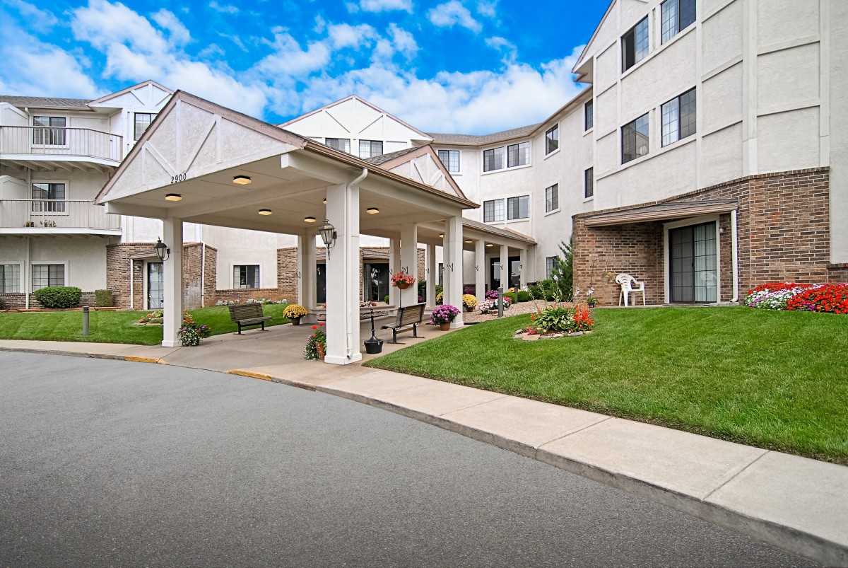 The image shows the entrance of a residential building with a driveway and landscaped surroundings.