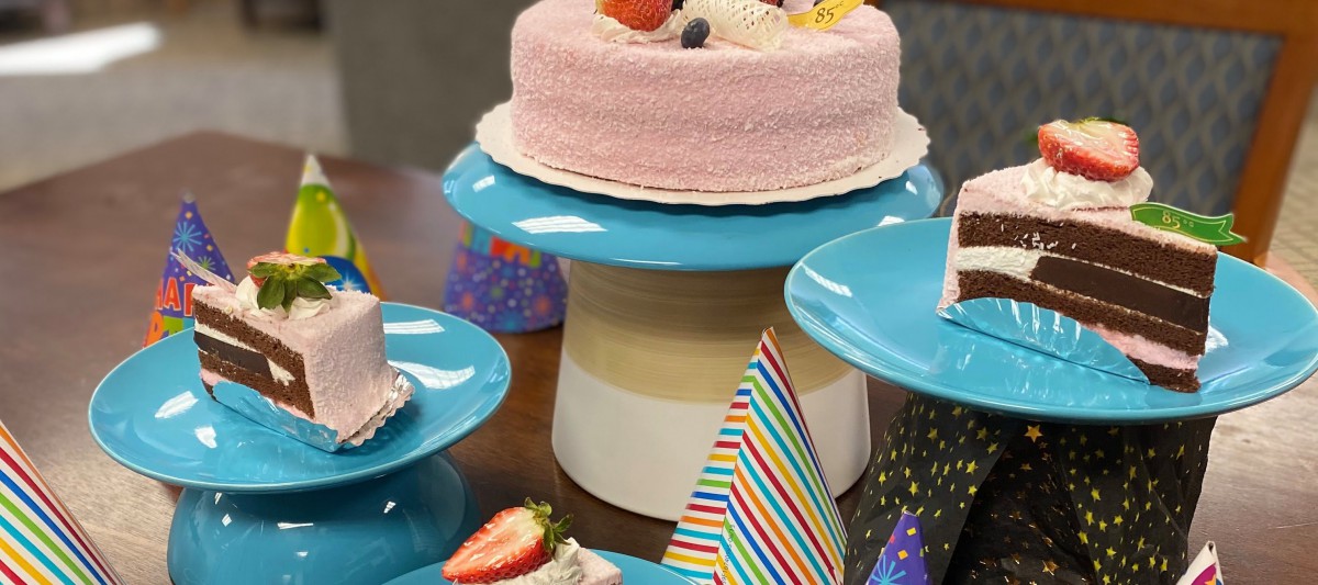A pink frosted cake with fruit toppings sits on a blue stand, surrounded by colorful party hats.
