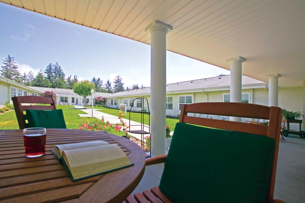 A peaceful outdoor patio area with a wooden table and chairs, featuring a book and overlooking a garden and buildings in the background.