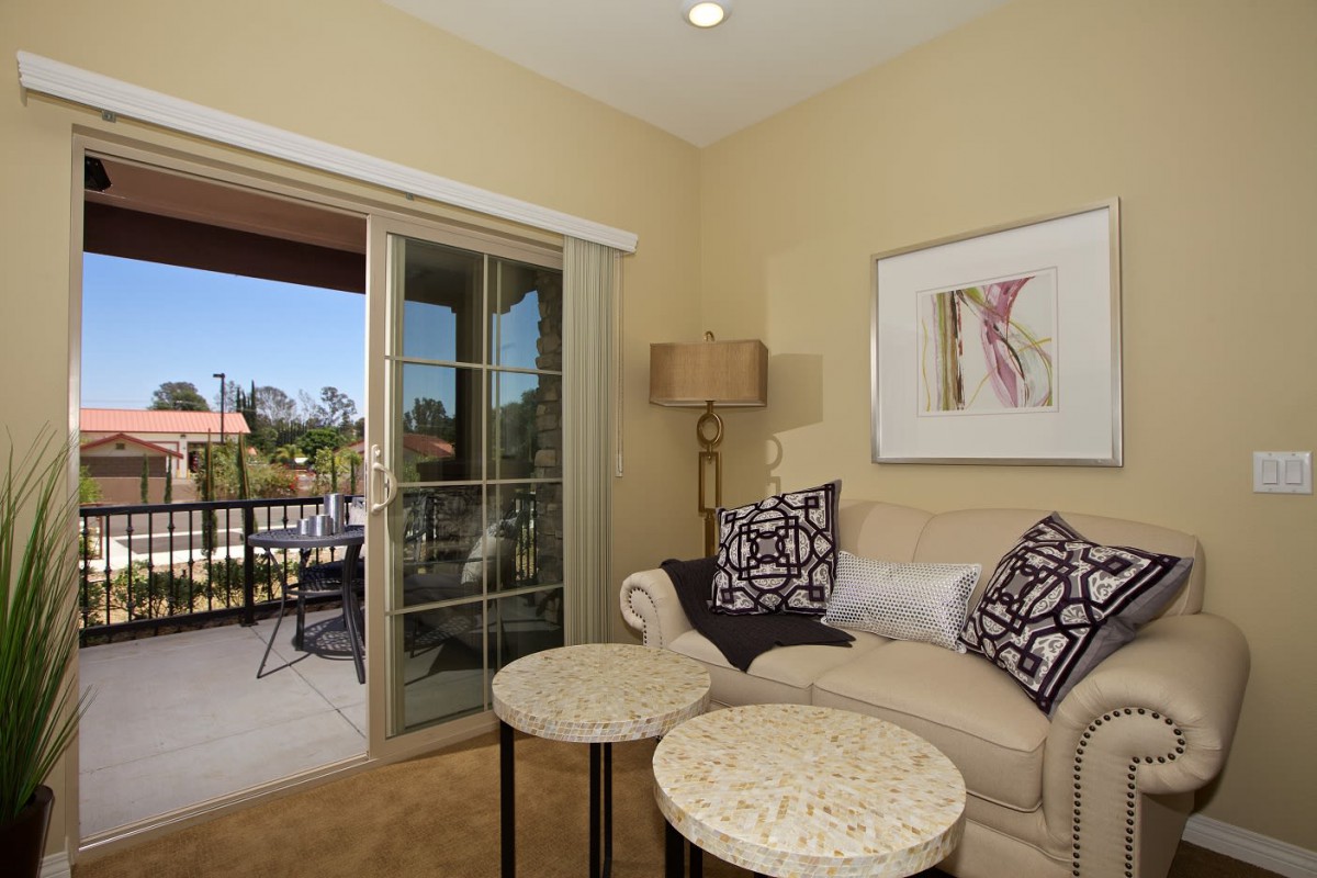 A cozy living room with a beige sofa, patterned pillows, and two small round coffee tables near a sliding glass door leading to a balcony.