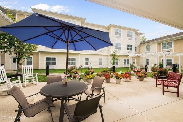 A sunny outdoor patio with a table, chairs, and a large blue umbrella, set against a backdrop of a multi-story residential building with a garden.