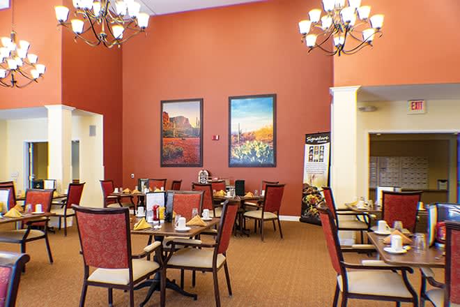 A cozy dining area featuring elegant chandeliers, warm red walls adorned with artistic paintings, and neatly arranged tables and chairs.