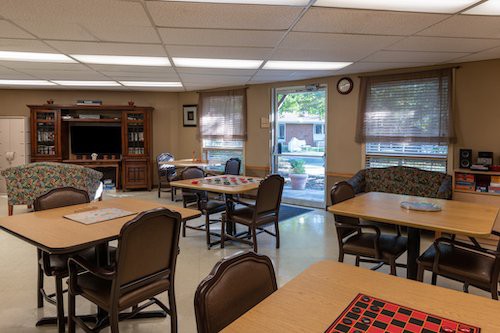 A recreational room with tables and chairs arranged for board games, featuring a TV stand and natural light from windows.