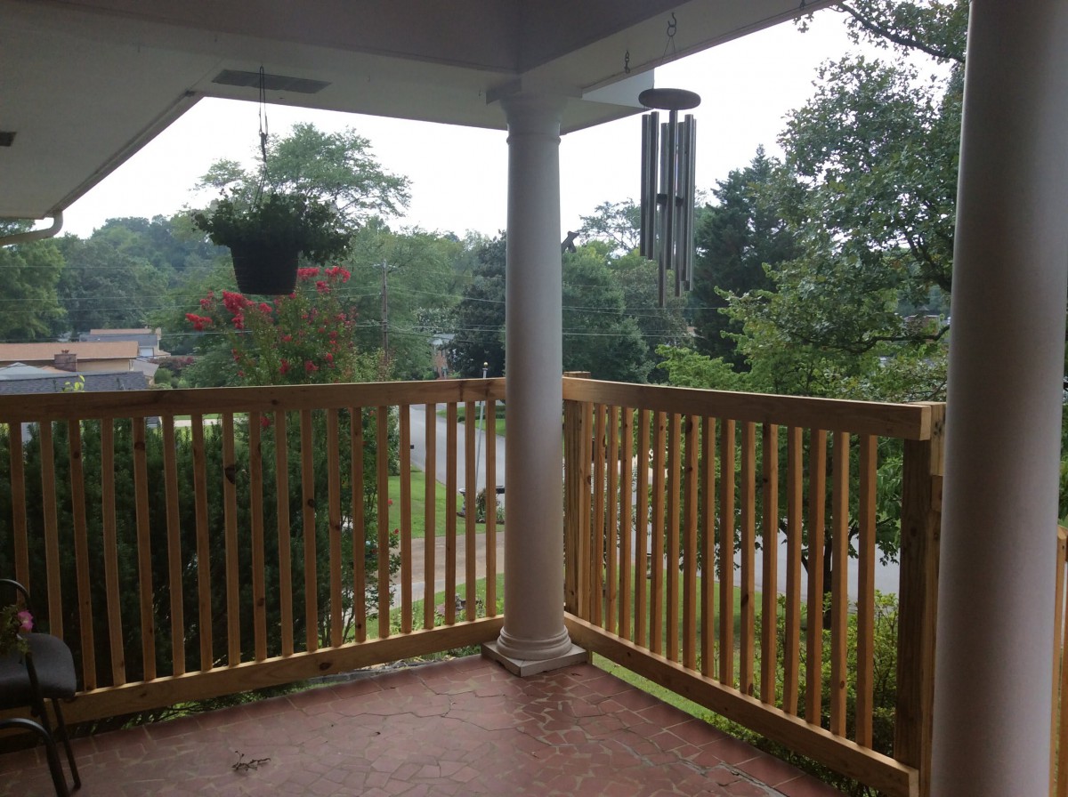A cozy balcony with wooden railings, featuring a hanging potted plant and a wind chime, overlooks trees on an overcast day.