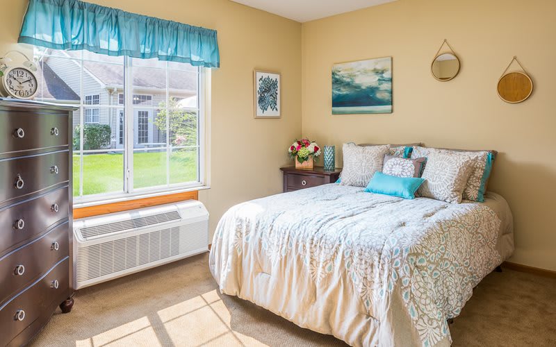 A cozy bedroom with a bed near a sunlit window, decorated with curtains and artwork on the walls.