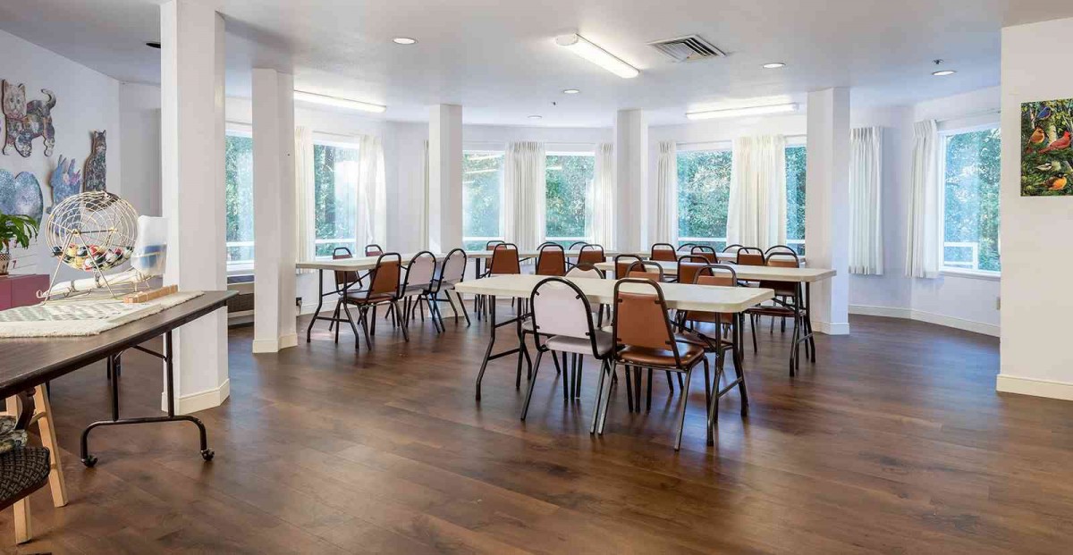 A bright room with multiple tables and chairs arranged on a wooden floor, surrounded by large windows.