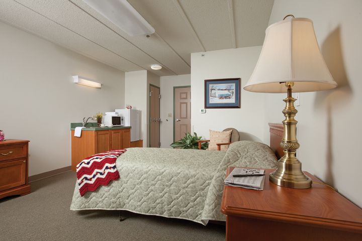 A cozy hotel room featuring a neatly made bed, bedside table with a lamp, and a small kitchenette in the corner.