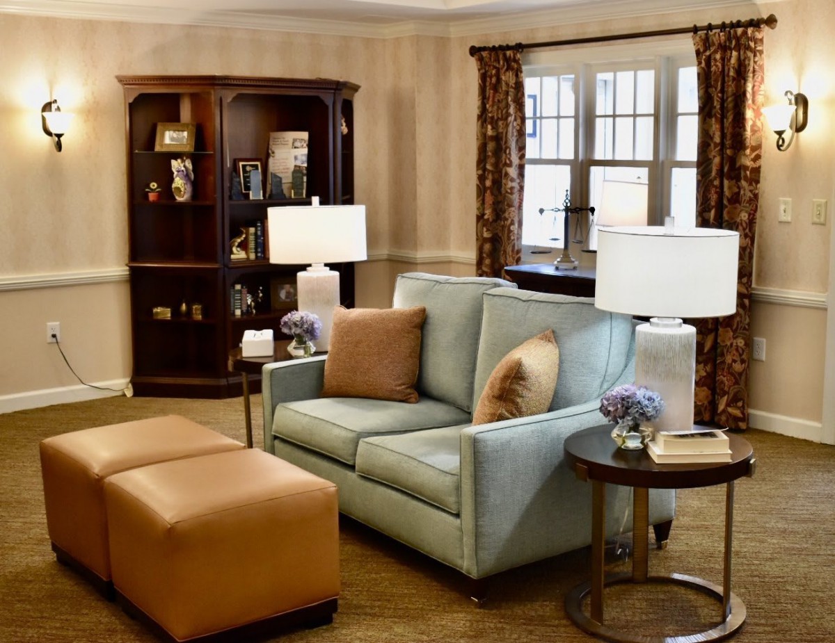 A cozy living room with a comfortable sofa, armchair, and ottoman, complemented by warm lighting from table lamps. A wooden bookshelf stands in the corner.