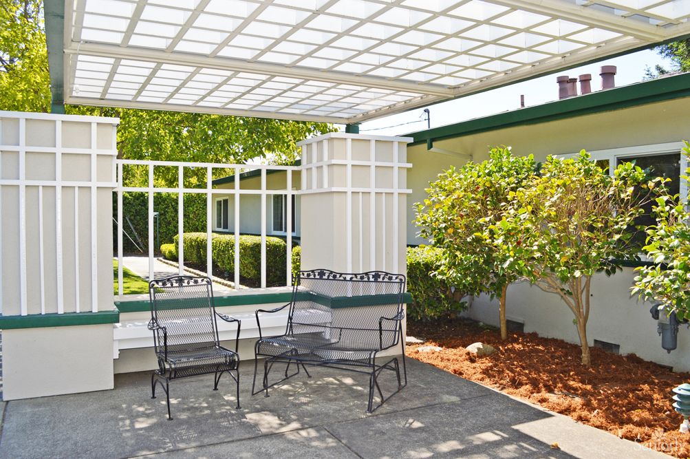 An outdoor patio with metal chairs under a pergola, surrounded by greenery.
