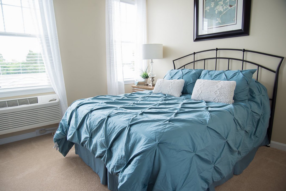 A cozy bedroom with a neatly made bed featuring blue bedding and pillows, complemented by natural light from the window.