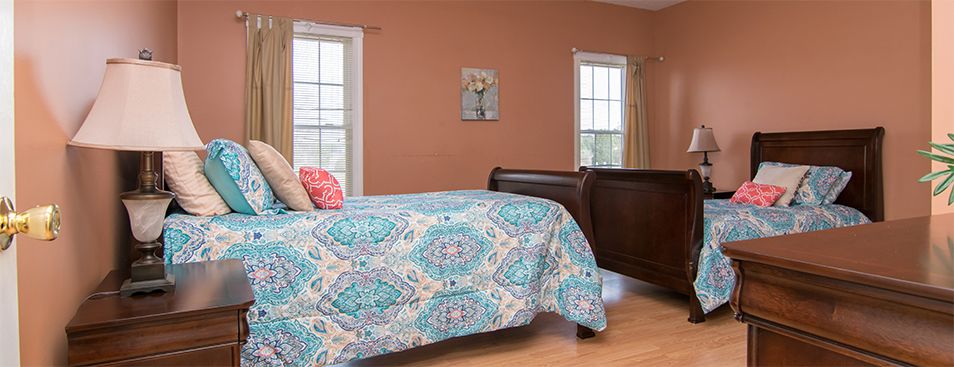 A cozy bedroom with a patterned quilt and warm-toned walls, featuring two windows and decorative wall art.
