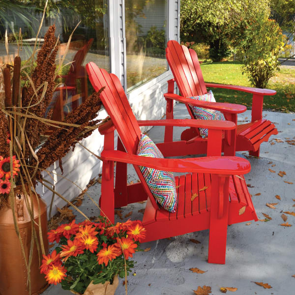 Two red Adirondack chairs on a patio surrounded by fall decorations and flowers.