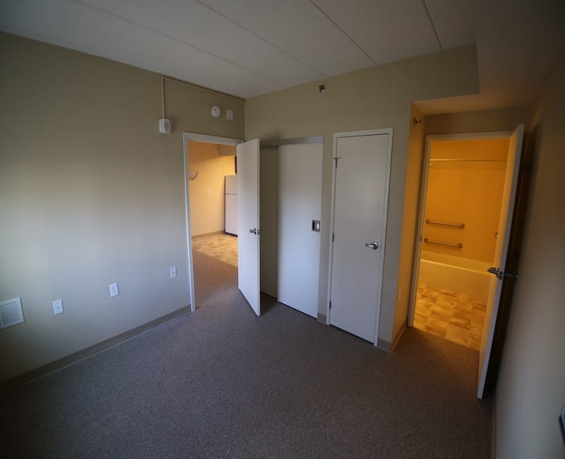 An empty room with carpeted floors, multiple doors, and a view into an adjoining hallway and bathroom.